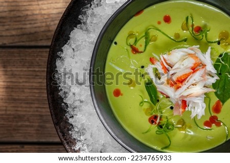 Broccoli and spinach cream soup with crab meat, herbs and balsamic sauce. Soup in a black ceramic plate on ice crumbs in a metal bowl. Dishes stand on a wooden background.