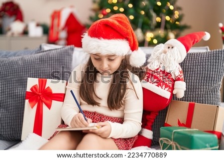 Adorable hispanic girl writing on notebook sitting on sofa by christmas tree at home