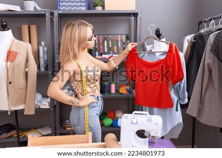 Young blonde woman tailor holding t shirt standing by manikin at atelier