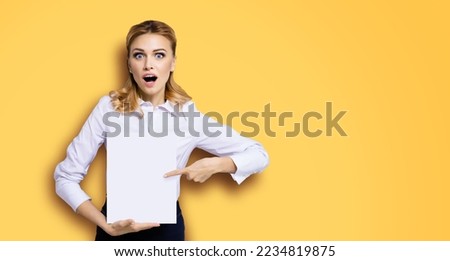 Very happy, excited surprised, astonished businesswoman with wide opened eyes, mouth, showing blank paper signboard. Success in business concept. Copy space place for text. Orange yellow background.