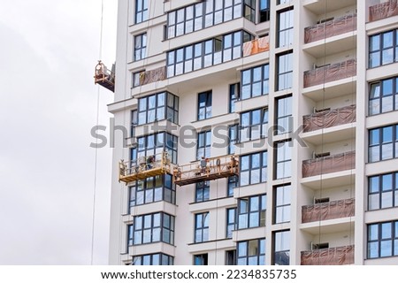 Workers in lift platform caulk, seal exterior glass window, maintain exterior glass facade. Men in lifting platform at height insulate and seal around building windows. Worker in suspended platform Royalty-Free Stock Photo #2234835735