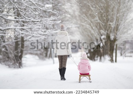 Young adult mother pulling wooden sledge with little girl on snow covered trail at park. Enjoying white, cold winter day. Spending time together in weekend. Back view.
