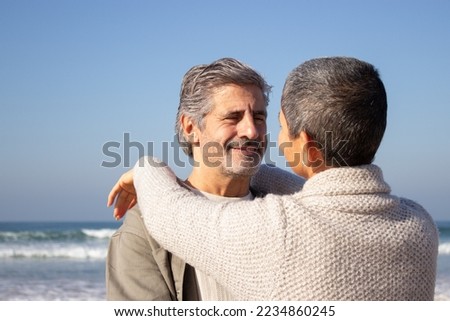 Handsome senior man embracing his wife and smiling while short-haired woman laying arms on mans shoulder. Middle-aged couple enjoying day at seashore. Sea background. Relationship, romance concept