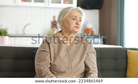 Unhappy and grumpy old woman is resting in her quiet home. Lonely old woman sitting alone on the sofa looking at the empty advertising space to the right of the screen.
