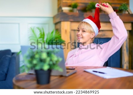 Photo of a young woman in a Santa hat in front of a laptop monitor in home interior. Online meeting with friends or relatives on the eve of Christmas or New Year.