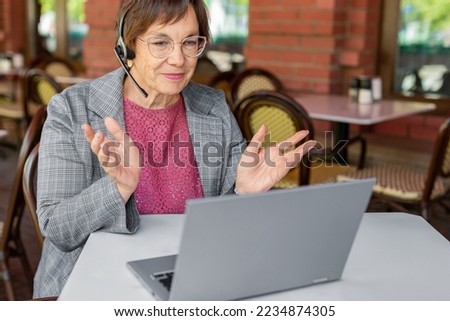 Close up photo of a senior woman during online video call. She i working in front of laptop monitor while sitting in summer cafe. Th concept of modern technology in every day life.