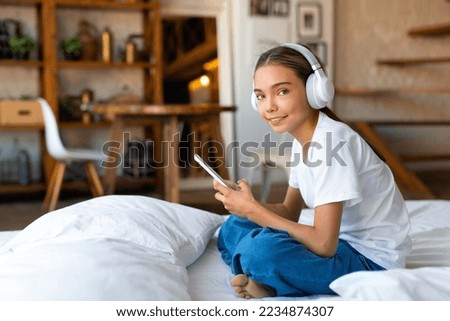 Teenager girl is sitting on the bed wearing headphones with a mobile phone. She listens to music through an online mobile application. The concept of children's dependence on mobile phones.