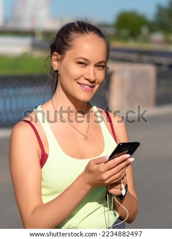 A young woman chooses music for her morning run. She is holding a smartphone with headphones, smiling and looking at the camera. Vertical photo.