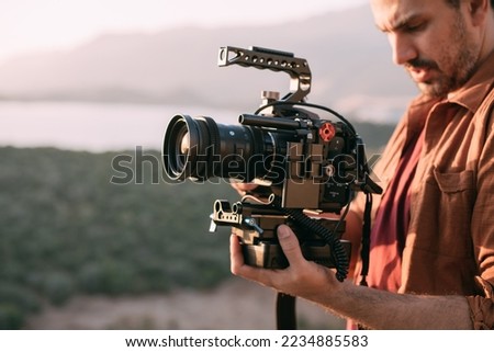 A young Caucasian man with a professional camera in his hands on a mountain by the sea.  The operator holds a video camera, shoots a sunset landscape, mountains, sea, setting sun Royalty-Free Stock Photo #2234885583
