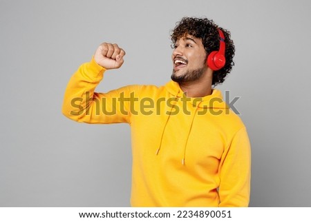 Young fun Indian man 20s he wearing casual yellow hoody headphones listen to music sing song in microphone at karaoke club isolated on plain grey background studio portrait. People lifestyle portrait