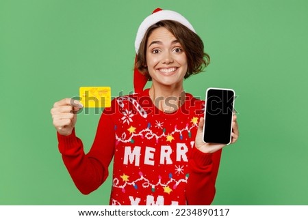 Merry young woman wears xmas sweater Santa hat posing hold blank screen mobile cell phone credit bank card shopping online isolated on plain pastel light green background Happy New Year 2023 concept