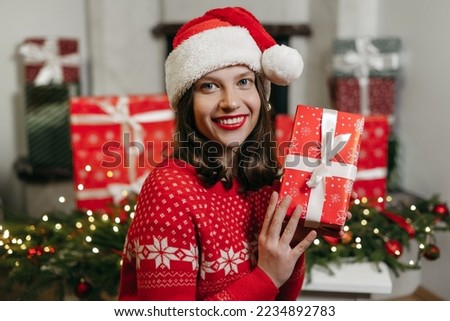 Portrait of a young beautiful happy woman in a Christmas hat, red sweater holding holiday gifts at home on background of bokeh garland lights, fireplace. New Year's mood, celebration. Smiled girl