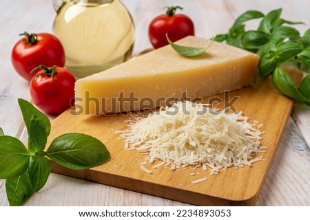 Parmesan and tomatoes on a cutting board. Italian grana padano cheese wedge and grated, red tomatoes, green basil herb and olive oil over white wooden table. Delicious hard cheese. Front view. Royalty-Free Stock Photo #2234893053
