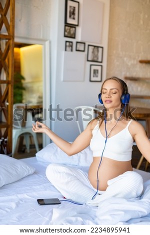 Health care concept - young pregnant woman sits on a bed wearing headphones and listens to music for meditation through an online smartphone application. Vertical photo.