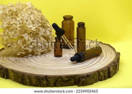 Mockup of brown glass vials with dropper lid on wooden board. Empty glass bottles with pipette, dried hydrangea flower on yellow background. Concept of medicine and face care. Selective focus.