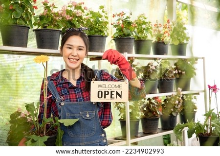 Portrait of beautiful smiling Asian woman looking at camera, holding open wooden sign board " Welcome we are OPEN in the greenhouse. Flowers shop and Store for sell plants