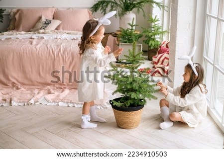 Two cheerful sisters decorate a Christmas tree in a pot in the early morning . kids are getting ready for the New year and Xmas . Girls in headbands with ears and in white dresses