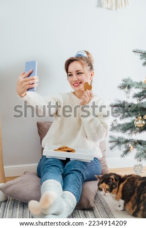 Young Woman in sweater taking selfie photo while relaxing with cat on floor cusions near potted christmas tree in modern Scandi interior home. Cozy winter holiday. Content creating. Selective focus