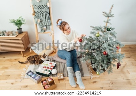 Young woman in cozy sweater petting her cat while decorating potted Christmas tree in light modern Scandinavian interior home. Eco-friendly winter holidays. Domestic pet at home. Selective focus