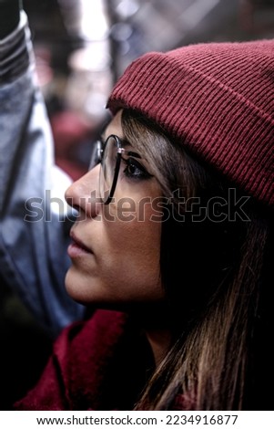 Close up picture of a hipster woman in magenta clothes checking the phone in the metro. She is standing and wear red winter clothes and denim jacket. Blurred background with people non recognizable