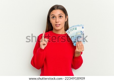 Young Indian woman holding a banknotes isolated on white background pointing upside with opened mouth.