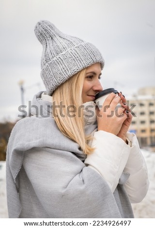 Young woman in beige fur coat, hat with pompon, scarf and white mittens holding steaming white cup of hot tea or coffee, outdoor in sunny winter day, close up 