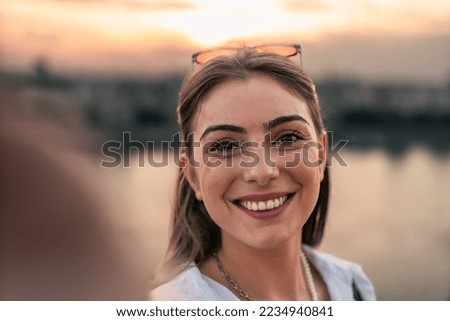 a portrait of beautiful girl tourist sitting on brick fence of old fortress. she is using her phone to taking photos of herself with city and river in background.