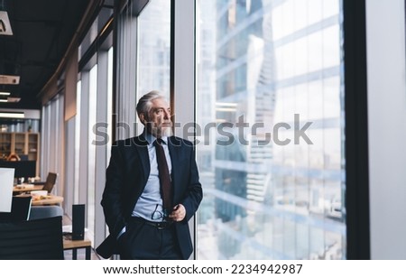 Pensive senior male CEO with gray hair in classy clothes holding eyeglasses and looking through window while working in modern office Royalty-Free Stock Photo #2234942987