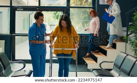 Asian patient using walking frame to walk in medical clinic, doing physiotherapy with nurse to recover and cure leg fracture. Person with physical impairment walking with stick in lobby.