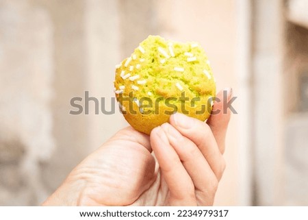 freshly backed pistachio biscuit with sugar sticks on top. Woman eats and hold bakery in hand outdoors. Traditional italian cookie in fingers with pastel colours street in background. Close up picture