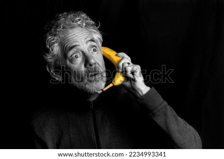 Humorous portrait of an attractive older Caucasian man with grey hair and beard, holding a banana like a telephone.  Appears suprised. Black and white photograph except for the yellow banana.
