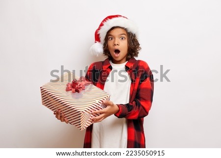 Cute teenager dressed in Santa Claus hat, holds a present and opened mouth in a shock and surprise, isolated over white wall