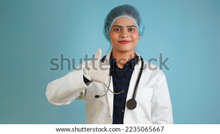 Portrait of a young female doctor gesturing thumbs up, Cheerful Asian Indian woman doctor in apron and stethoscope isolated over blue studio background