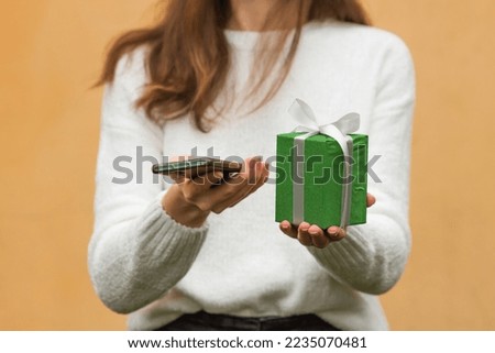 Woman's hands holds smartphone and green gift box with bow. Female in white sweater using mobile app and shopping online