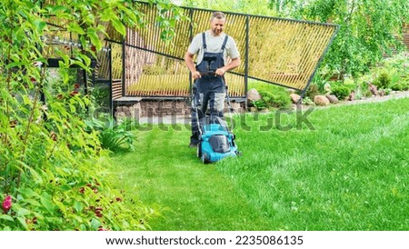 A man mows the grass with an electric lawn mower on a hot summer day. Gardener with a lawn mower in a beautiful landscape design. A hired worker tends the garden and does housework. Gardener services. Royalty-Free Stock Photo #2235086135