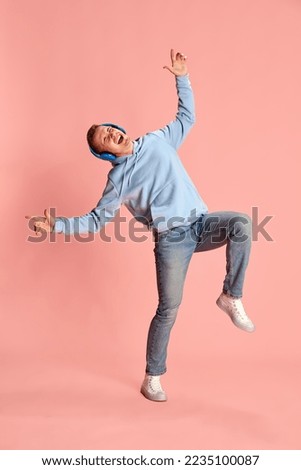 Portrait of young emotional man in hoodie and jeans listening to music in headphones and dancing isolated over pink background. Concept of youth, lifestyle, music, fashion, emotions, facial expression