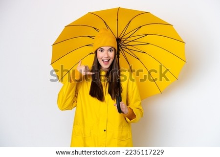 Young Brazilian woman with rainproof coat and umbrella isolated on white background making phone gesture. Call me back sign