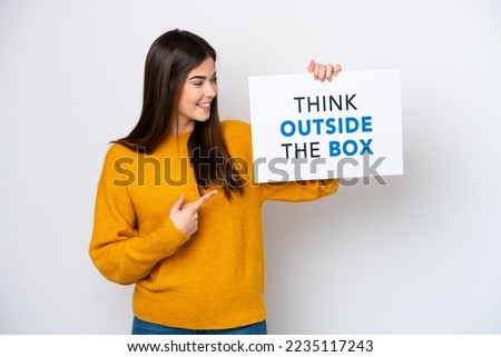 Young Brazilian woman isolated on white background holding a placard with text Think Outside The Box and  pointing it