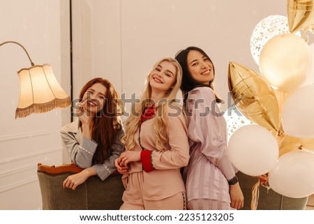Smiling three young interracial girls posing for camera spending time at sleepover. Blonde, brunette and redheads wear pajamas indoors with balls. Lifestyle concept