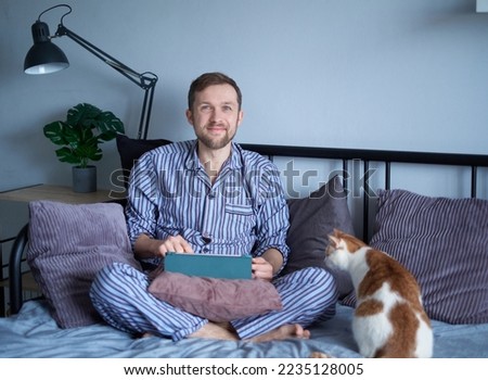 Male using tablet computer wearing sleep wear sitting on bed at home. Making purchases online or surfing the internet with tabby cat nearby. High quality image Royalty-Free Stock Photo #2235128005