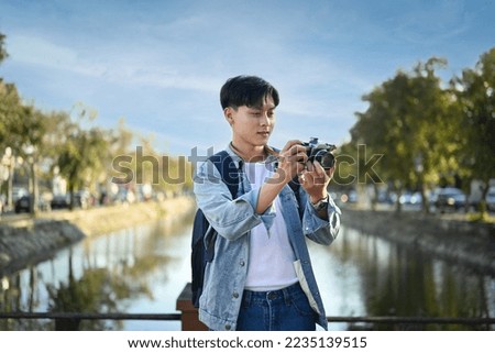 Asian man traveller with backpack taking photo with digital camera while standing on a bridge over a canal