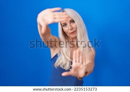 Caucasian woman standing over blue background doing frame using hands palms and fingers, camera perspective 