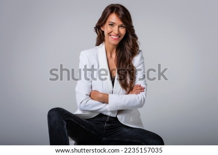 Attractive middle aged woman with toothy smile wearing blazer while sitting at isolated dark background. Copy space. Studio shot.