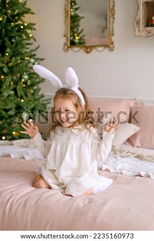 A beautiful cute girl with rabbit ears in a nightgown is sitting on the bed and laughing merrily spreading her arms to the sides. Xmas ornate decorated Christmas tree in the bedroom