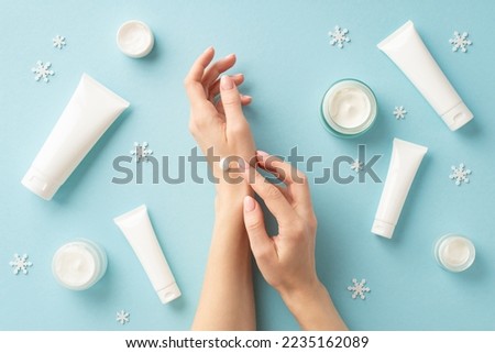 Winter skin care cosmetics concept. First person top view photo of woman applying hand cream over jars tubes without label and snowflakes on isolated light blue background with empty space