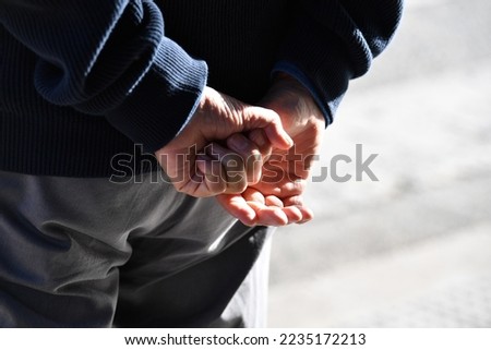 An elderly man's hand holds his hands behind his back.