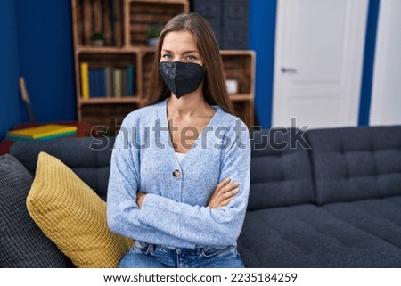 Young woman wearing medical mask sitting on sofa with arms crossed gesture at home