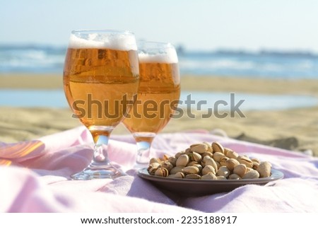 Glasses of cold beer and pistachios on sandy beach near sea, closeup