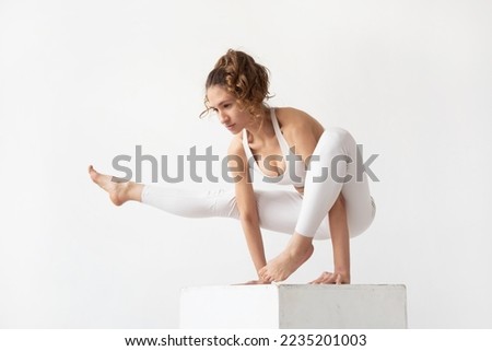 Young woman leading a healthy lifestyle and practicing yoga, performs the exercise eka pada bhujapidasana, handstand on a white cube, trains in white sportswear on a light background