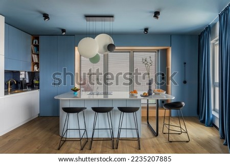 Interior design of kitchen space with marble island, black chockers, modern lamp, wooden wall, blue kitchen furnitures, vase with dried flowers and personal accessories. Home decor. Template. 